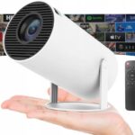 Baltas projektorius Reach HY300 laikomas delne su juodu nuotolinio valdymo pulteliu ir rodomas fone